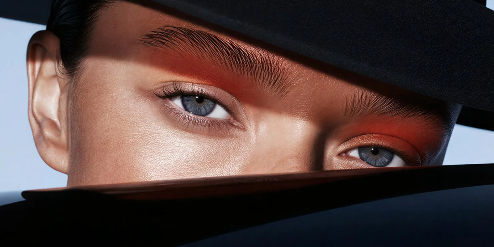 Close-up of a woman's blue eyes with orange eyeshadow under a black hat brim, makeup detail that is better filmed with real hands