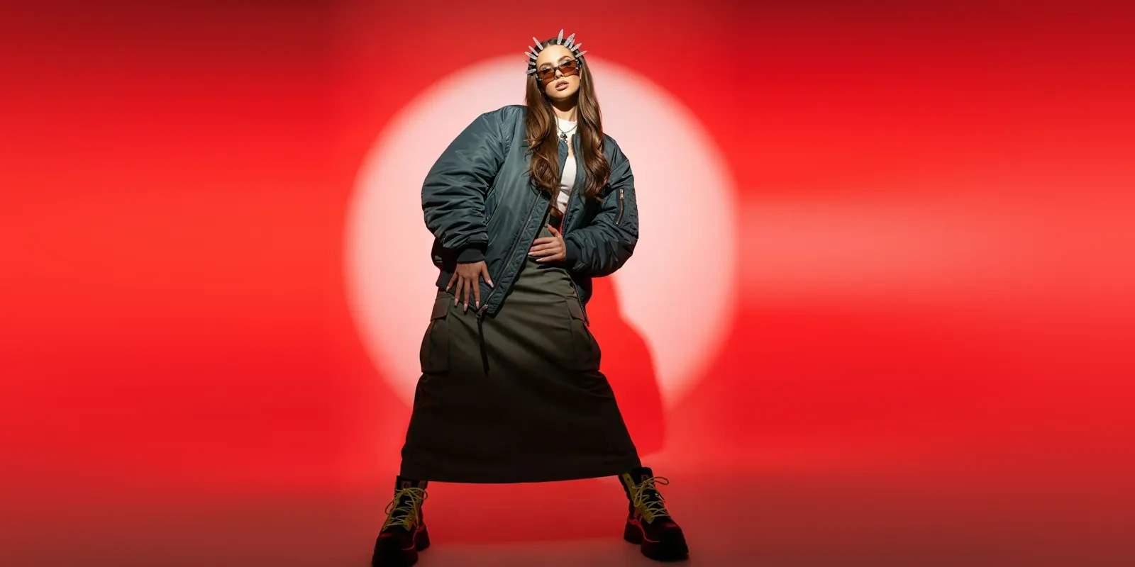 Woman in a green bomber jacket and boots poses under a spotlight before a red backdrop, a styled shoot that still suits a photographer