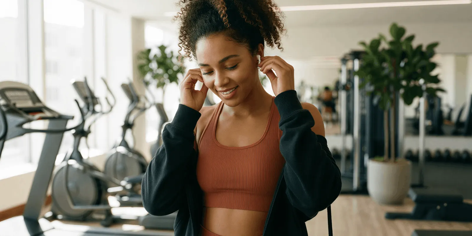 Woman in a rust sports top and black jacket puts in earbuds on a gym floor, a setting that shows where a product is used and promises no result