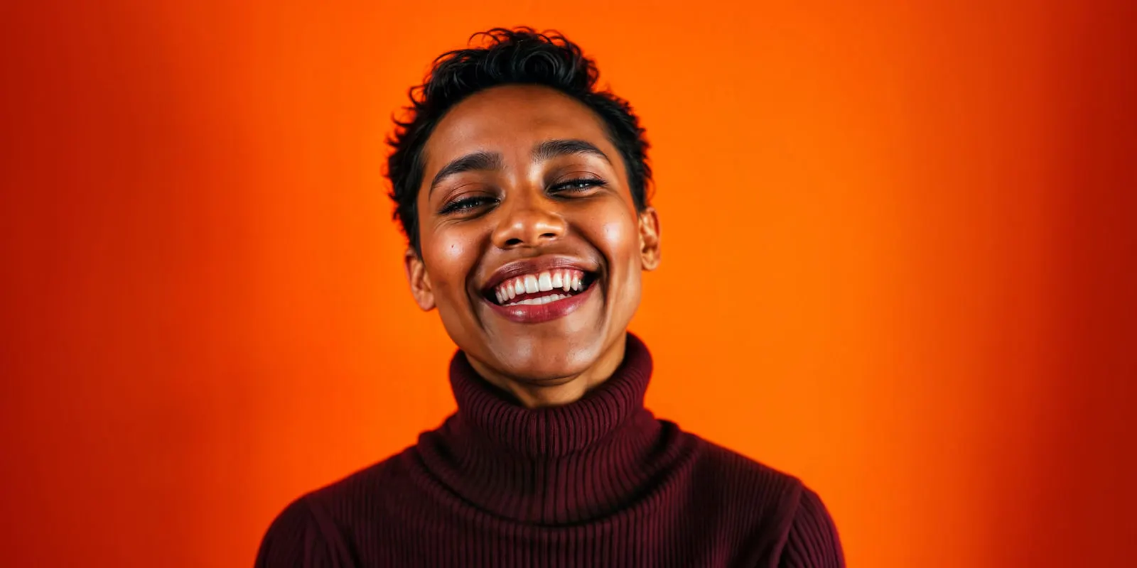 Person in a burgundy knit turtleneck laughing toward the camera against an orange backdrop, the on-model frame a fashion drop needs