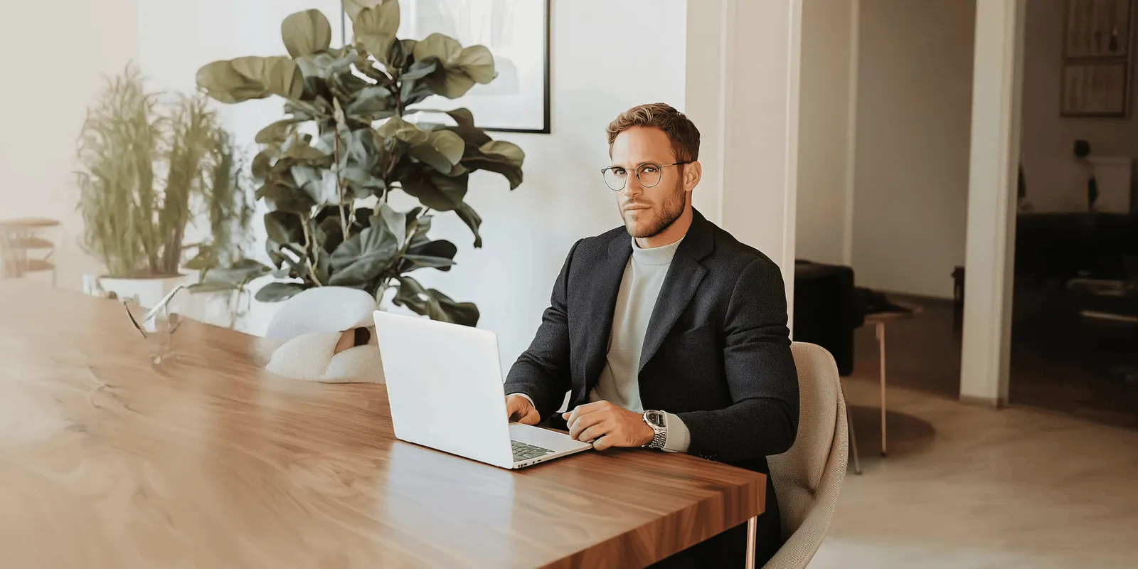 Man in a blazer and glasses at a wooden desk with an open laptop and large plants behind him, sorting a folder of product photos before a batch