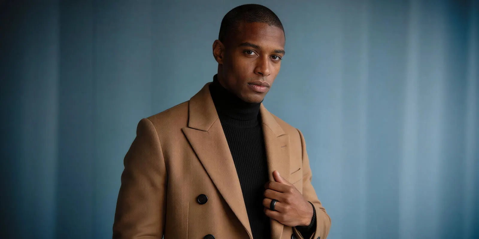 Man in a camel double-breasted coat over a black turtleneck against a blue-gray backdrop, the kind of frame a coat needs