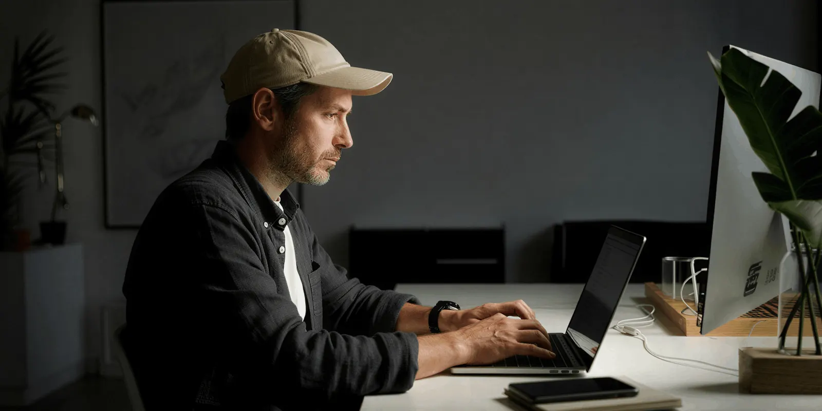 A man in a cap checks work on a laptop at a white desk, the final review before edited images go live