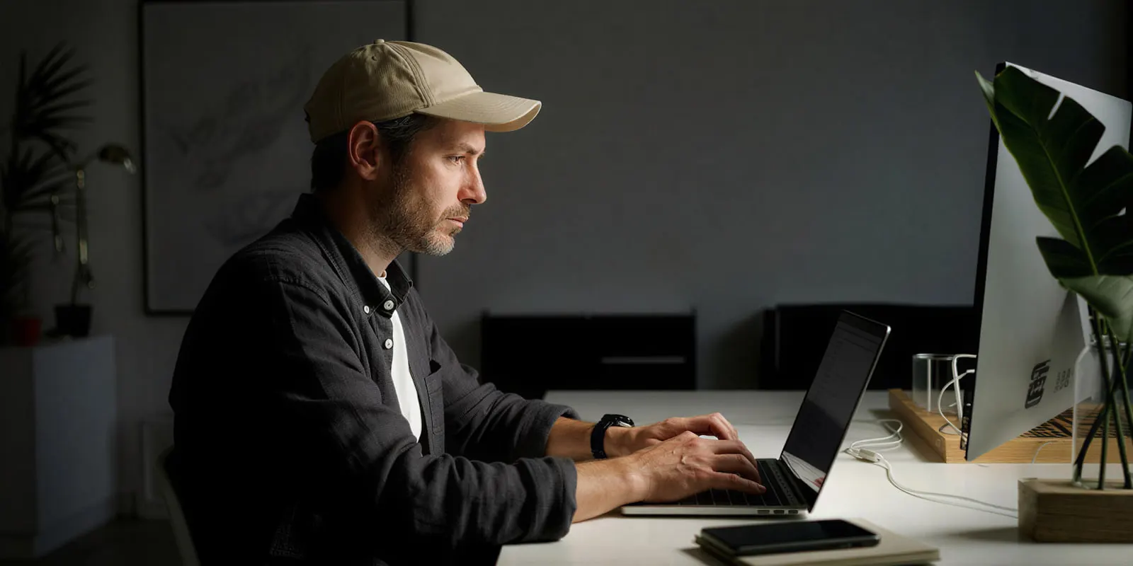 A man in a cap works at a laptop beside a large monitor, reading each result closely before it goes live