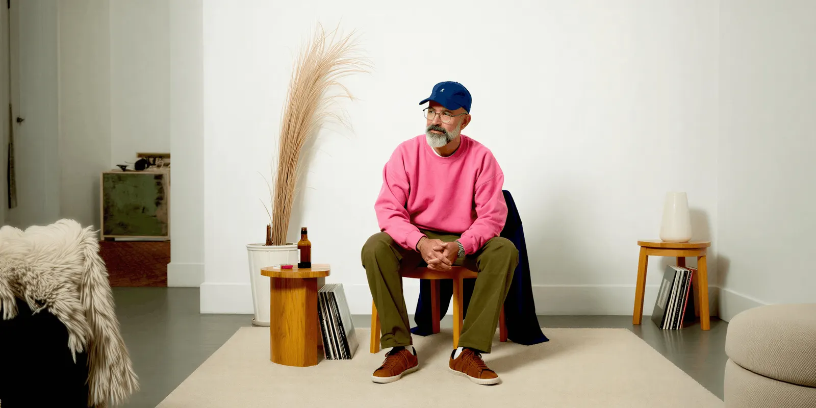 Man with a gray beard in a pink sweater, blue cap and olive trousers on a stool in a white room, a worn cap that shows fit