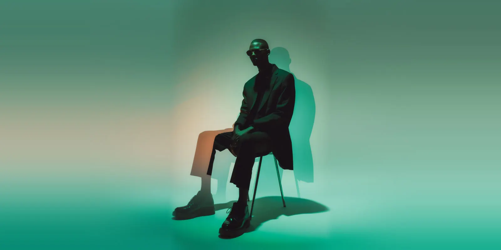 Man in a dark suit seated on a chair in a green-lit studio, the seated presenter shot that avatar tools set out to imitate