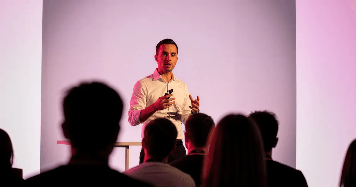 Man in a white shirt holding a clicker as he speaks to a seated audience, the meeting where a pilot project needs a clear result