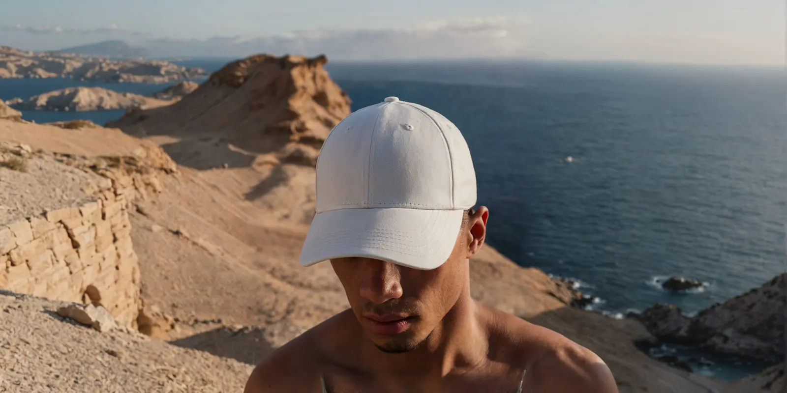 Man in a white cap on a coastal cliff above the sea in low evening sun, the kind of location a swim line is shot in