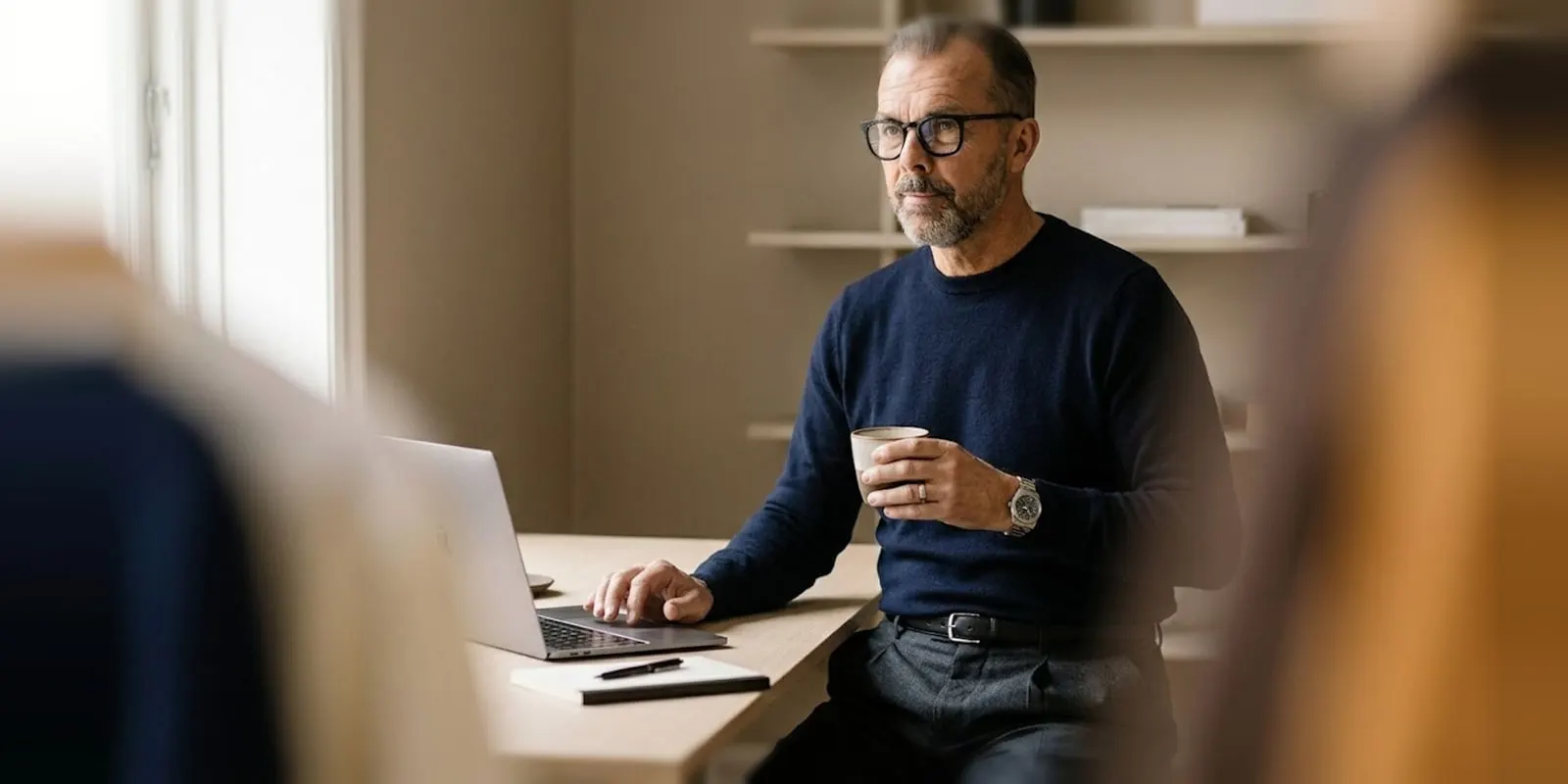 Man in a navy sweater holds a coffee at his desk beside a laptop and a pen, comparing plan prices before he picks one