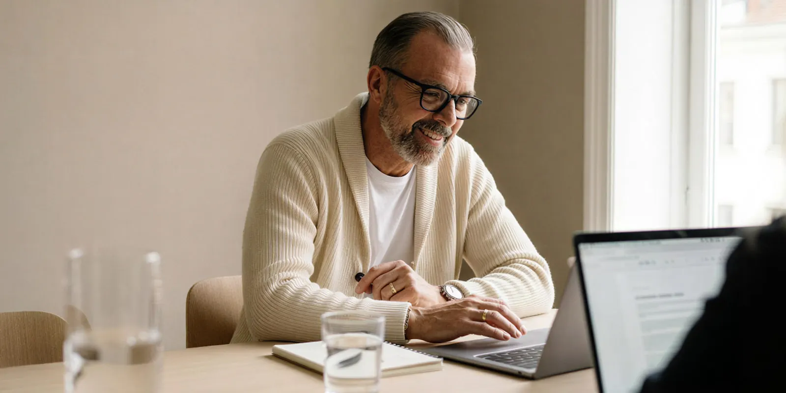 An older man in glasses and a cream cardigan uses a laptop with a notebook beside him, reading a model license before a campaign
