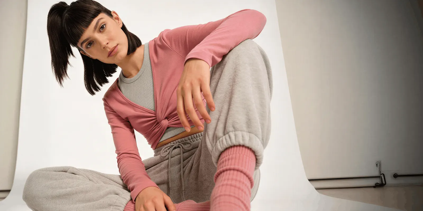 Model in a pink top and gray joggers seated on a white paper sweep, the plain set a catalog shoot uses for every product