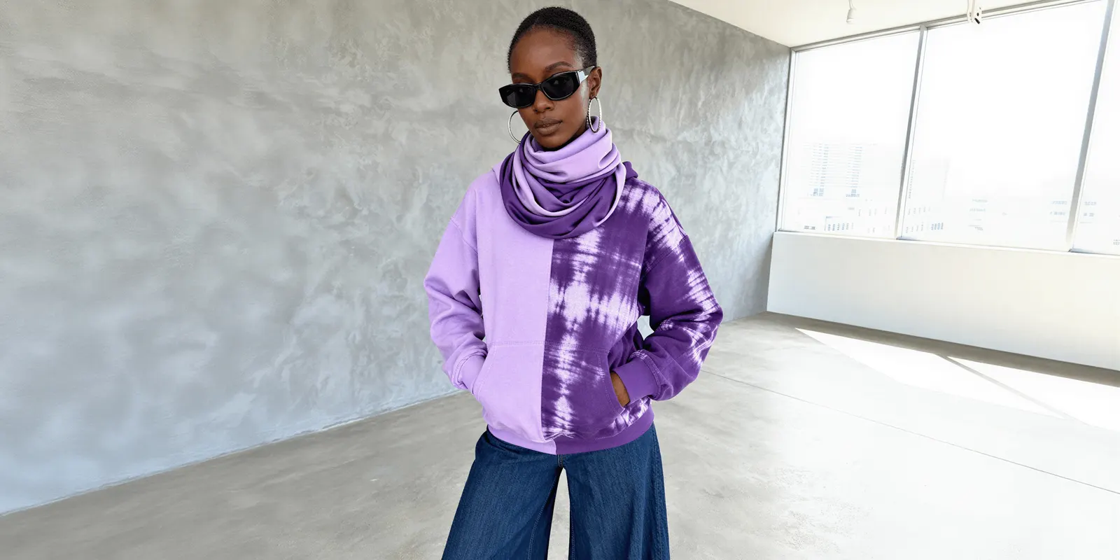 Model in a half lilac, half purple tie-dye hoodie against a grey wall, a fabric pattern that is hard for an image model to keep