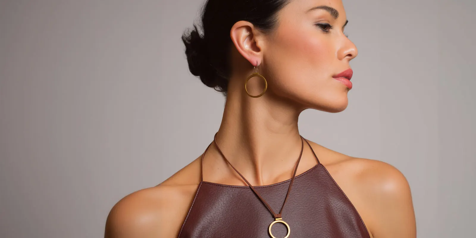 Woman in a brown leather halter top wearing a round pendant necklace and hoop earring, a necklace that reads in one frame