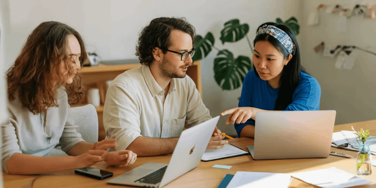 Three colleagues sit at a table and point at two laptop screens, comparing what they see the way a team checks results against a written standard
