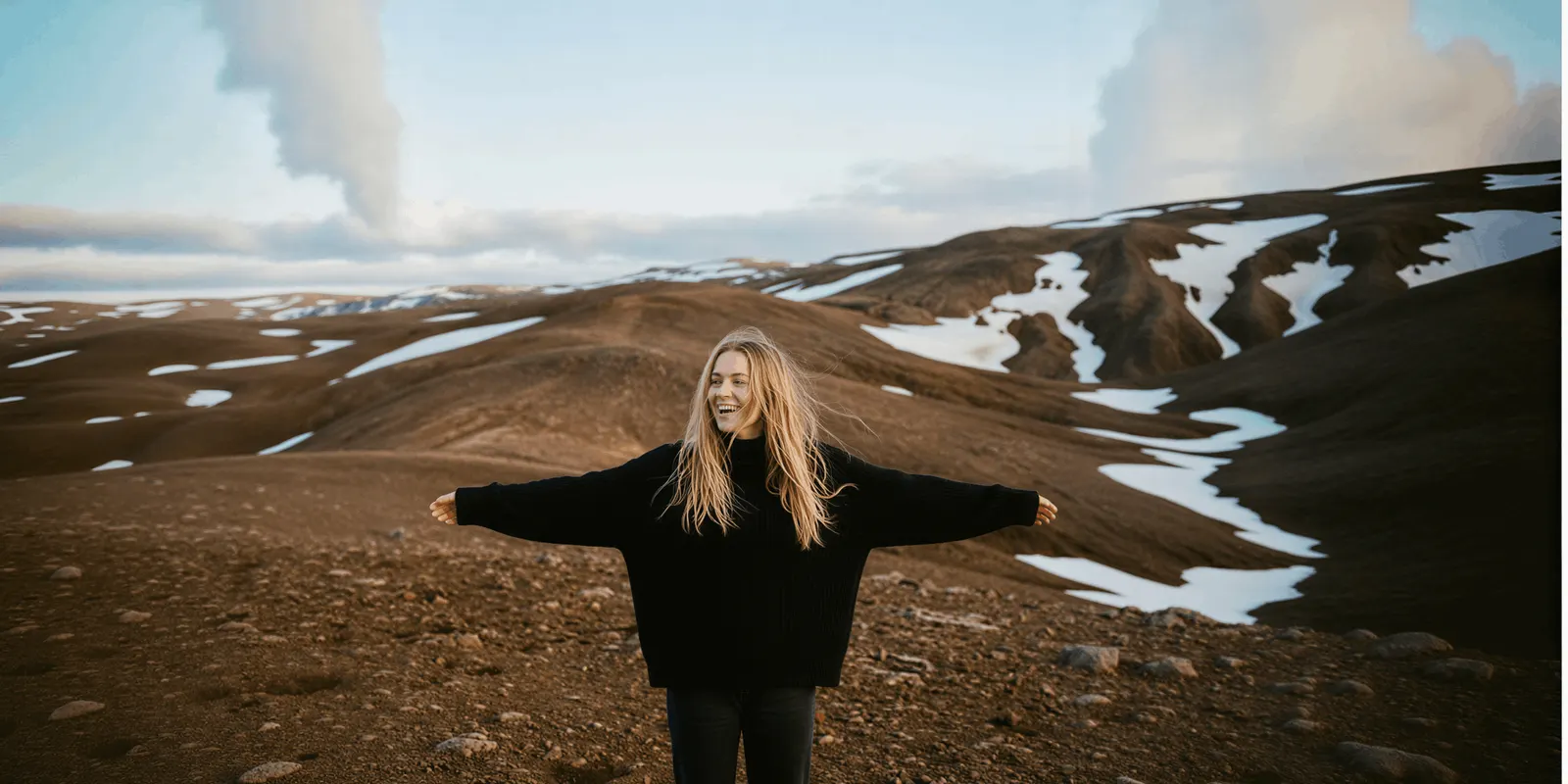 Woman laughing with arms out on a brown hillside with snow patches, the kind of real destination footage a travel ad has to show