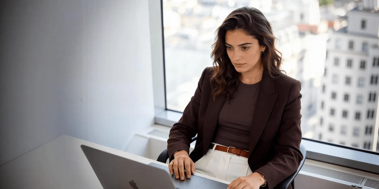 Woman in a brown blazer works at a laptop by a window over the city, weighing a design tool against an ad tool