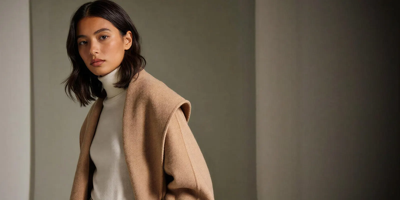 A woman in a camel coat over a cream turtleneck stands against a muted gray wall, one of many looks a single sample has to supply