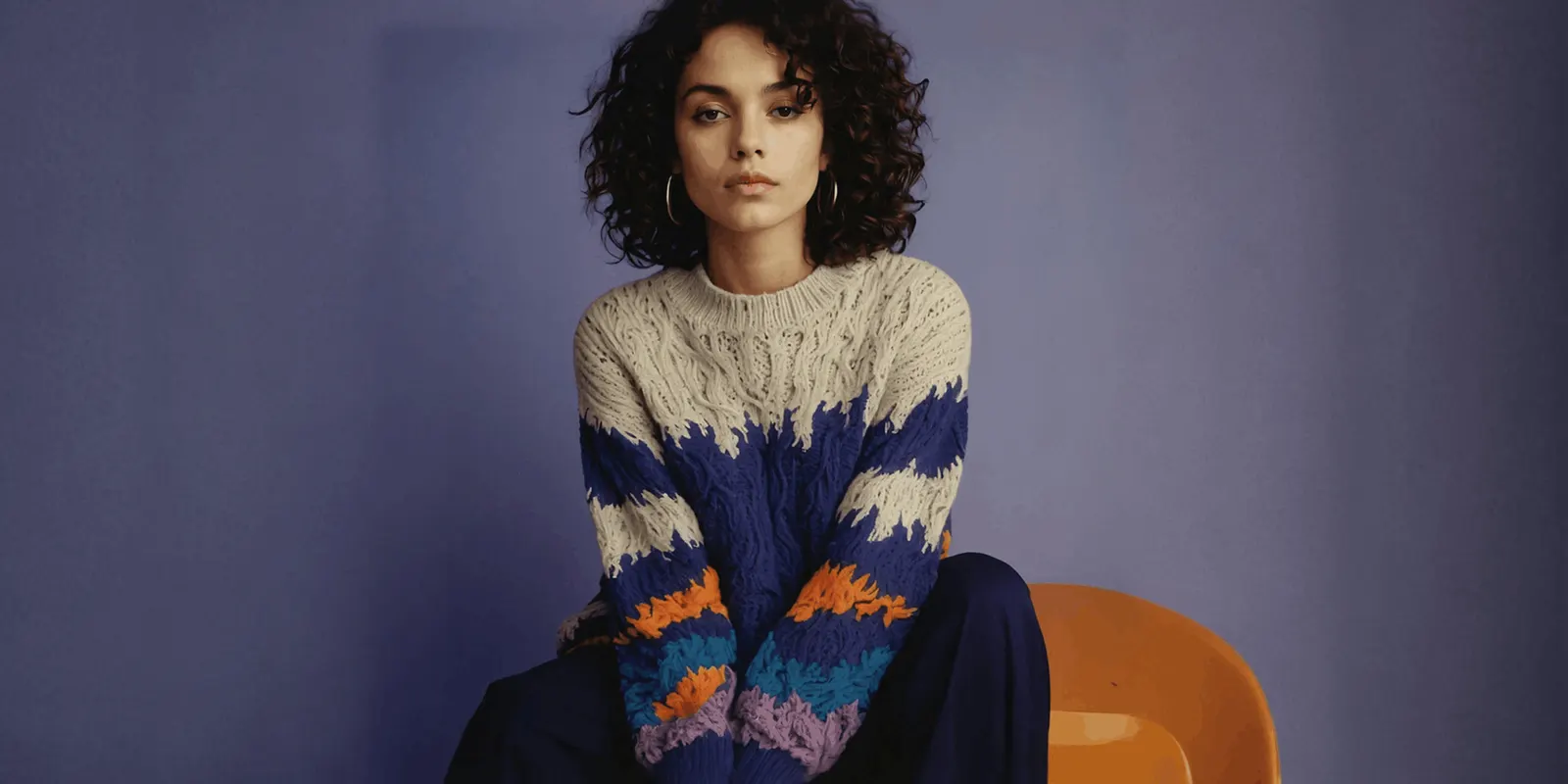 Woman in a cream, blue and orange knit sweater sits on an orange chair against a purple wall, one color version of a garment that needs its own image