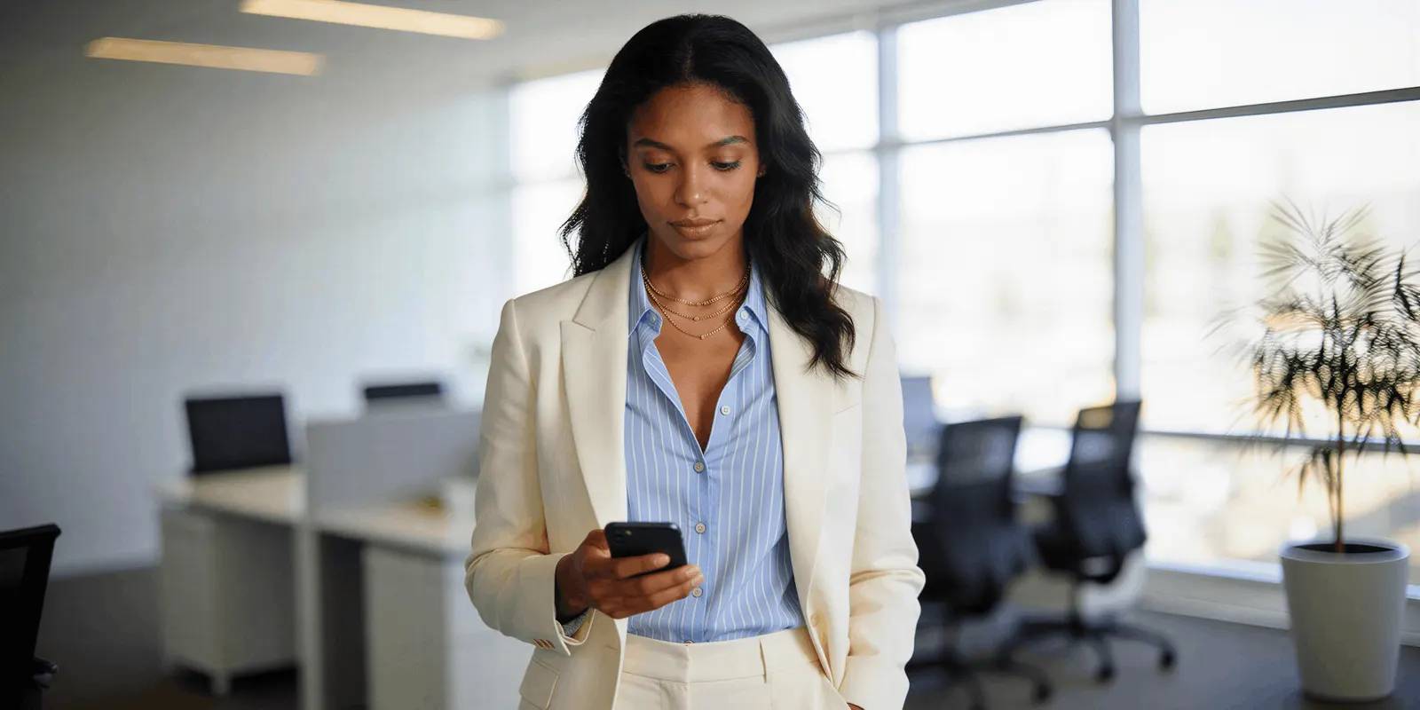 Woman in a cream blazer checking an app on her phone in an open office, the kind of real screen use an app ad shows