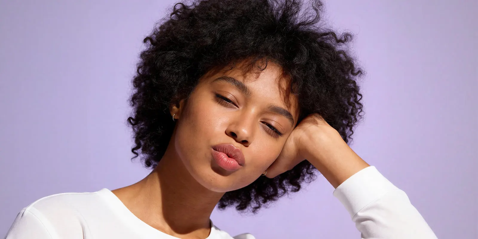 Woman with curly black hair in a white top rests her head on her hand against a lilac backdrop, a close crop where skin and hands must hold at zoom