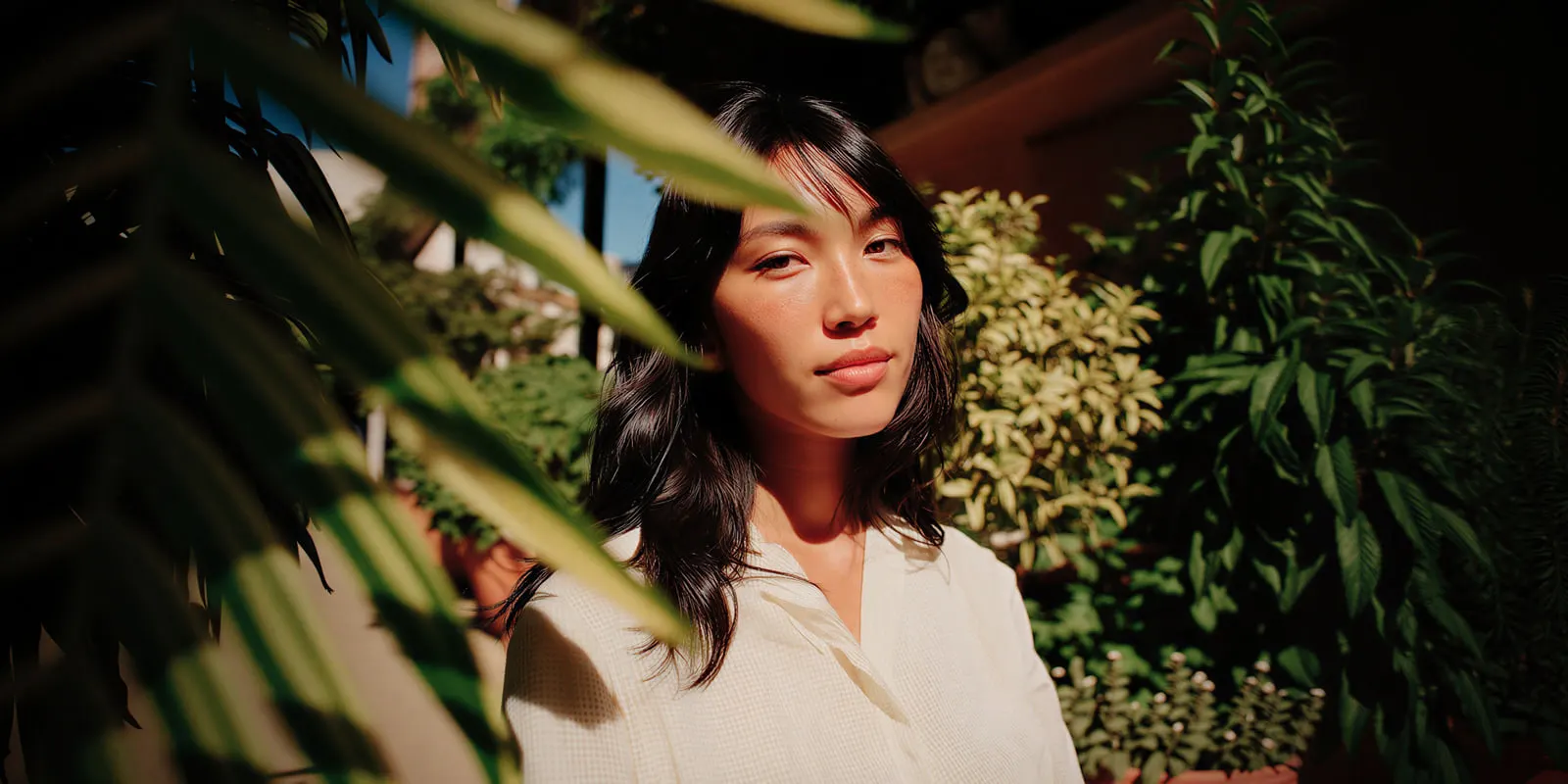 Woman in a cream shirt framed by green leaves in a sunlit garden, the kind of plant setting that reads as an ingredient claim