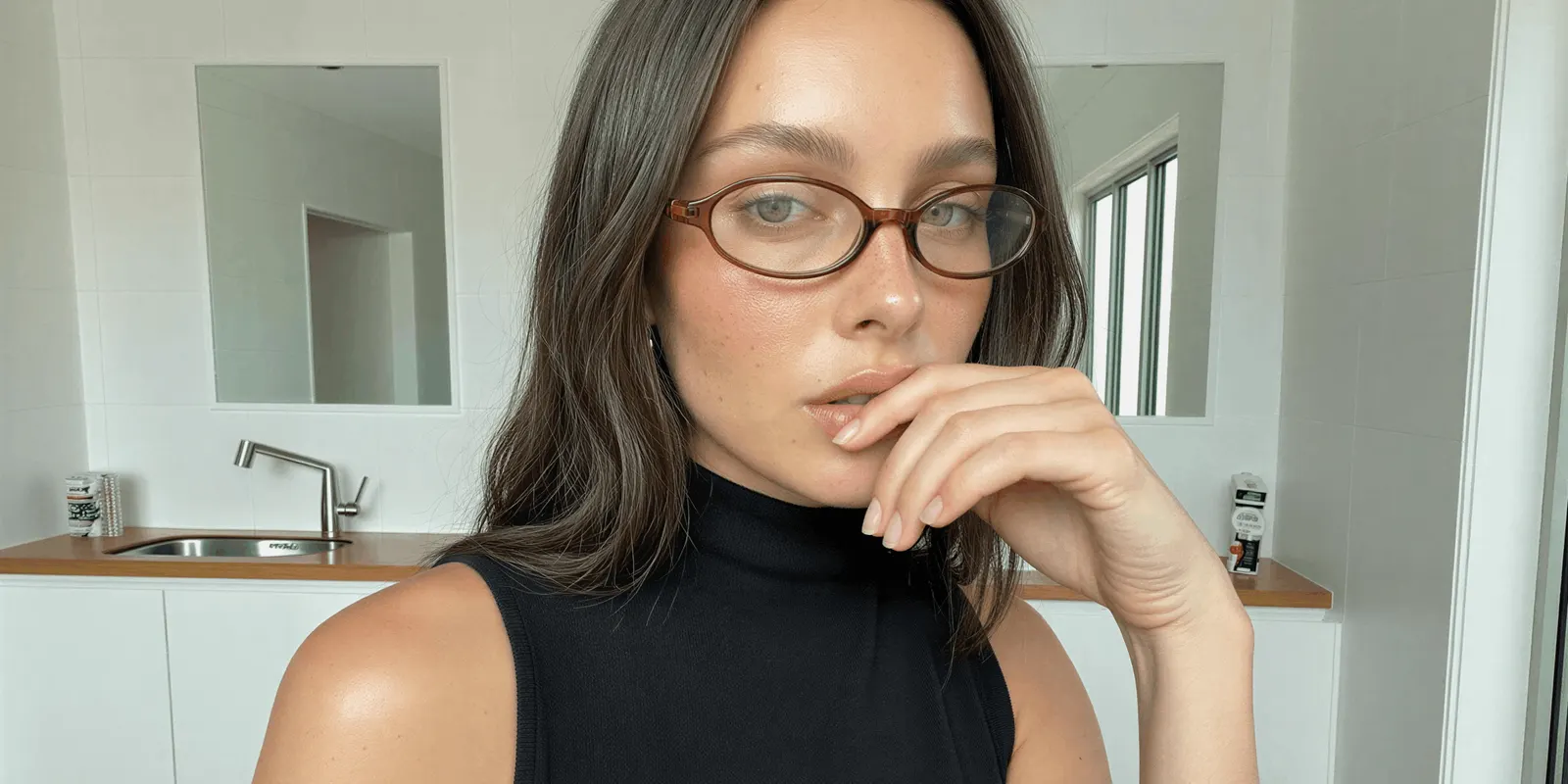 Close-up of a woman in thin oval glasses, hand at her lips, with visible pores and freckles: the skin texture a believable makeup ad keeps
