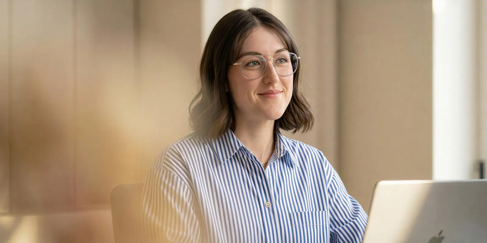Woman in glasses and a striped shirt at a laptop in a warm office, the kind of desk where one creator video is priced by its deliverable