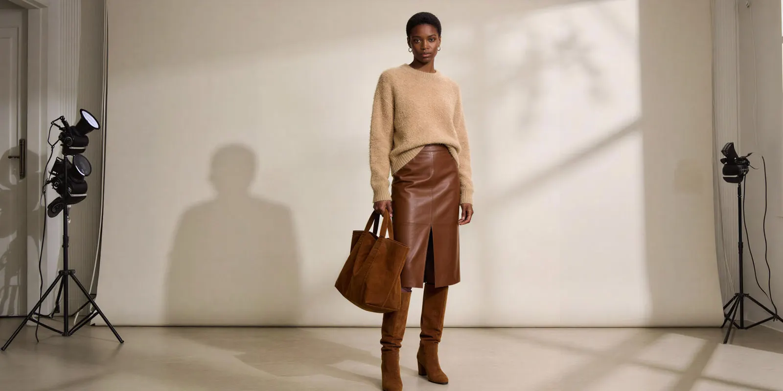 Woman in a camel knit sweater, leather skirt and boots holding a tote bag between studio light stands, the drop photography to fix first