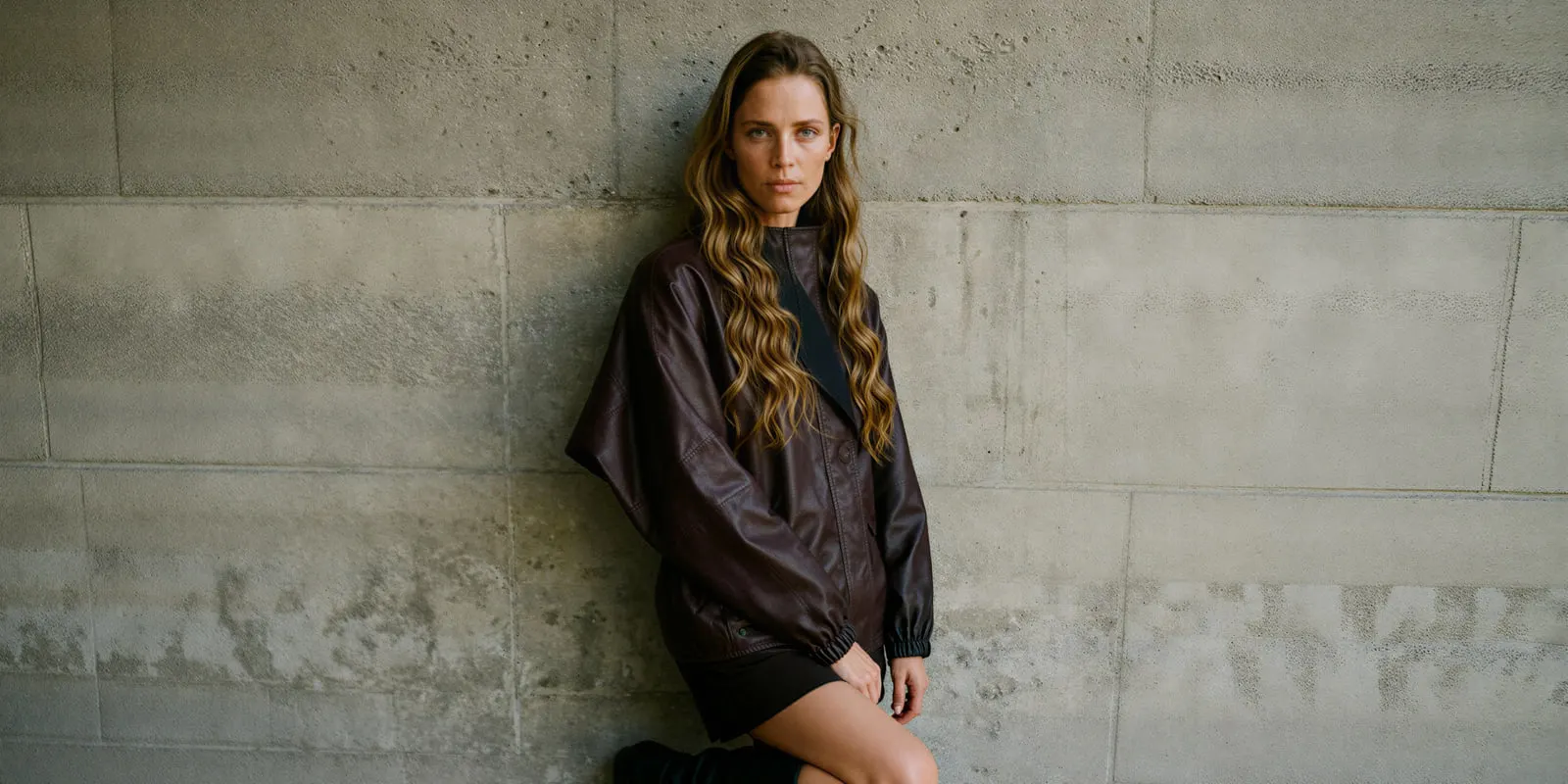 Woman in a leather jacket crouches against a concrete wall, the way weight and pose change how leather sits on a body