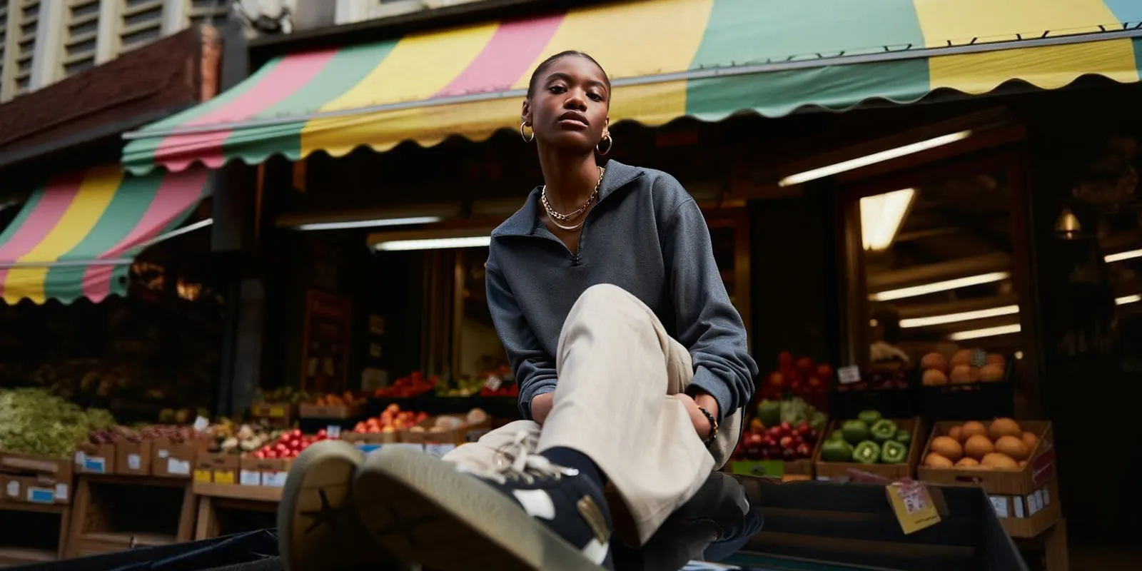Woman in a gray half-zip top and cream trousers sits under a striped awning at a fruit market, the everyday shopper fashion retail serves