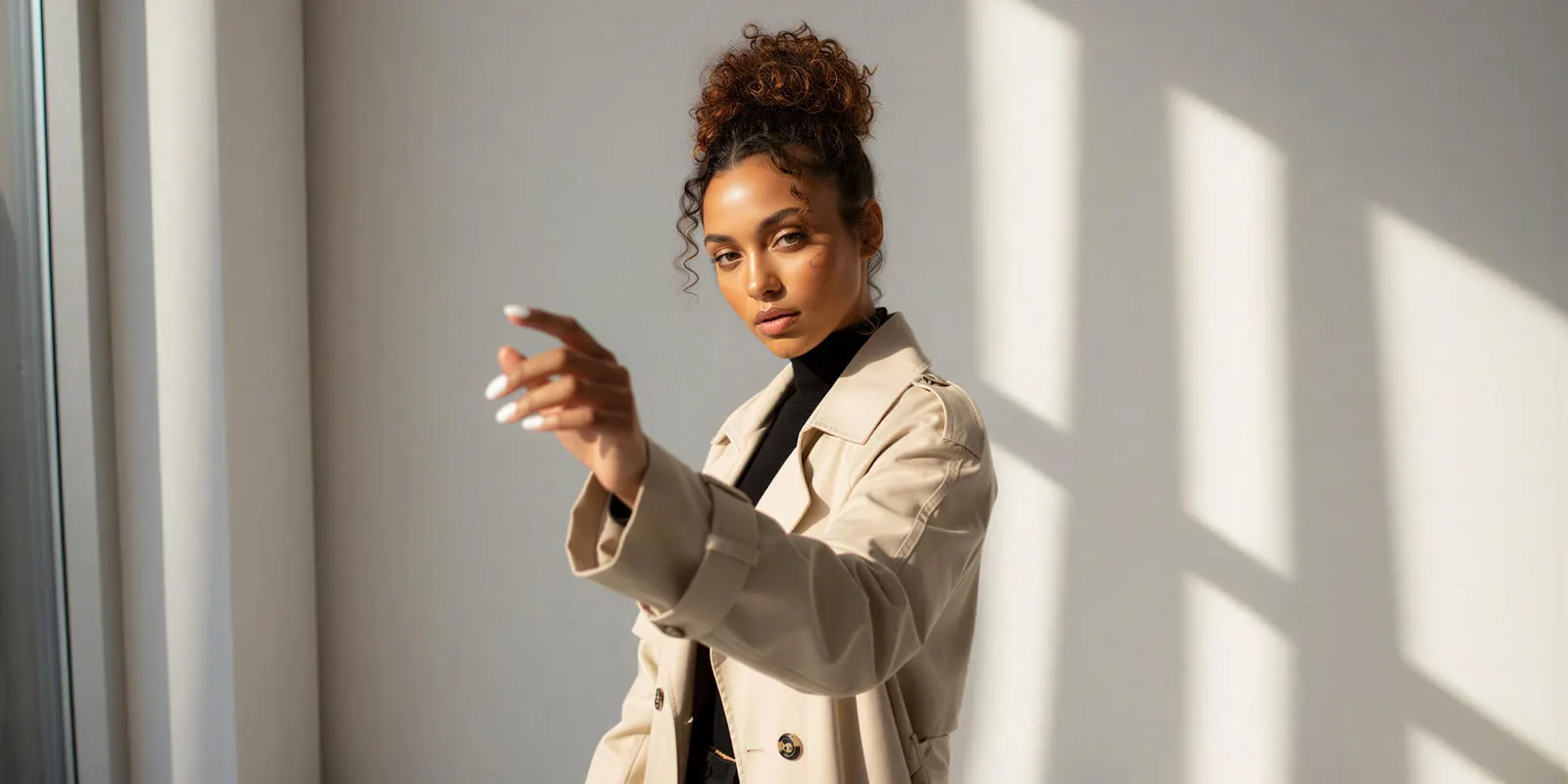 Woman in a beige trench coat extends a hand against a sunlit wall, the model and light a set keeps as the camera moves