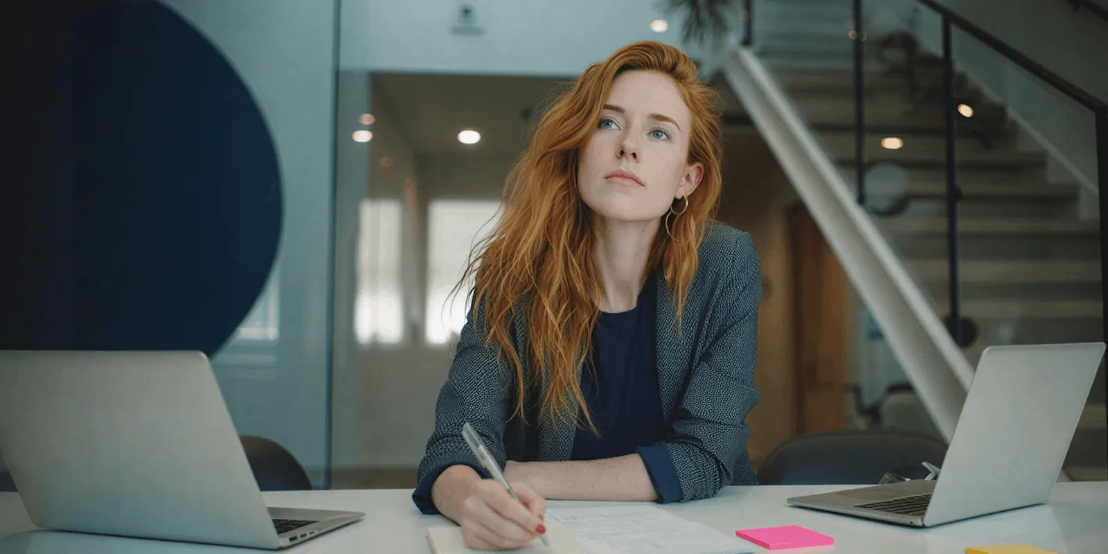 Woman writes notes on paper between two open laptops in an office, the side-by-side read a license and a set of terms need