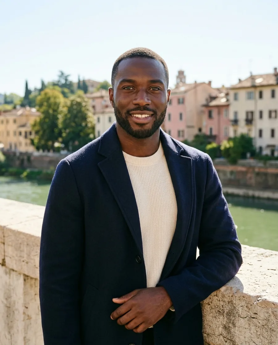 A smiling man with a short beard in a navy wool coat and a cream sweater on a sunny stone bridge in an old town, made with AI